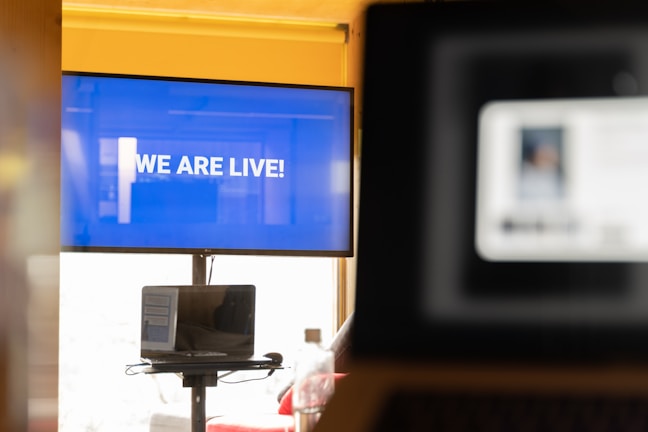 A large screen displays the message 'WE ARE LIVE!' in bold white lettering against a blue background. In front of the screen, there is a laptop on a stand showing an out-of-focus presentation slide. Another blurred screen is visible in the foreground, suggesting a busy digital or virtual environment.