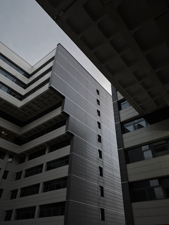 A modern building with a geometric design is depicted, characterized by sleek lines and a facade made of metal panels and glass. The angled perspective highlights the architectural structure against a cloudy sky, creating a sense of depth and symmetry.
