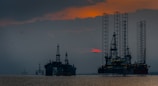 Silhouettes of offshore oil rigs set against a hazy sunset sky. The structures are positioned in the calm sea, with the orange and red hues of the sun partly obscured by clouds. Several birds are flying in the sky, adding to the serene yet industrial scene.