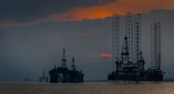 Silhouettes of offshore oil rigs set against a hazy sunset sky. The structures are positioned in the calm sea, with the orange and red hues of the sun partly obscured by clouds. Several birds are flying in the sky, adding to the serene yet industrial scene.