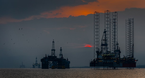 Silhouettes of offshore oil rigs set against a hazy sunset sky. The structures are positioned in the calm sea, with the orange and red hues of the sun partly obscured by clouds. Several birds are flying in the sky, adding to the serene yet industrial scene.