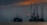 Silhouettes of offshore oil rigs set against a hazy sunset sky. The structures are positioned in the calm sea, with the orange and red hues of the sun partly obscured by clouds. Several birds are flying in the sky, adding to the serene yet industrial scene.
