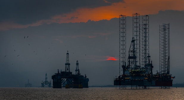 Silhouettes of offshore oil rigs set against a hazy sunset sky. The structures are positioned in the calm sea, with the orange and red hues of the sun partly obscured by clouds. Several birds are flying in the sky, adding to the serene yet industrial scene.
