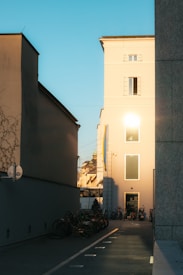The image captures a narrow alleyway between buildings, with bicycles lined up on one side. Sunlight reflects off the beige facade of one building, casting a warm glow. The sky is clear with a light blue hue, and there's a hint of urban architecture in the background.
