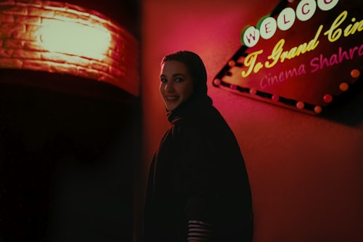 A woman is smiling and standing in front of a warmly lit cinema sign that reads 'Welcome To Grand Cinema Shahr'. The sign is adorned with colorful lights against a red and pink backdrop.