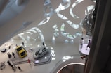 An interior view of a modern, futuristic exhibition space with a high ceiling featuring abstract, organic shapes. Two cars are displayed on platforms, one yellow and one silver, surrounded by people observing. A bicycle is also on display nearby. On the right, a glass elevator with people inside is visible. The space is well-lit with natural light pouring in through large glass windows.