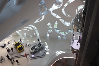 An interior view of a modern, futuristic exhibition space with a high ceiling featuring abstract, organic shapes. Two cars are displayed on platforms, one yellow and one silver, surrounded by people observing. A bicycle is also on display nearby. On the right, a glass elevator with people inside is visible. The space is well-lit with natural light pouring in through large glass windows.