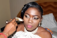 A woman is having makeup applied, possibly in preparation for a special event. She is wearing elegant jewelry and a white garment, indicative of a formal occasion. Her expression appears focused and serene as she looks slightly away from the camera. The background includes a bed, suggesting this moment is captured in a personal or intimate setting.