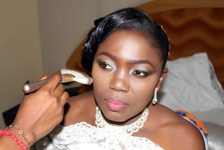 A woman is having makeup applied, possibly in preparation for a special event. She is wearing elegant jewelry and a white garment, indicative of a formal occasion. Her expression appears focused and serene as she looks slightly away from the camera. The background includes a bed, suggesting this moment is captured in a personal or intimate setting.