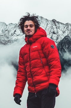 A person wearing a bright red puffer jacket stands in the foreground with misty mountains in the background. The scene is set in a cold, mountainous environment.