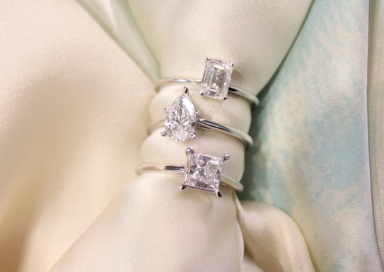 Three diamond rings with different cuts, including cushion, pear, and emerald, are elegantly displayed on a soft, light-colored fabric backdrop.