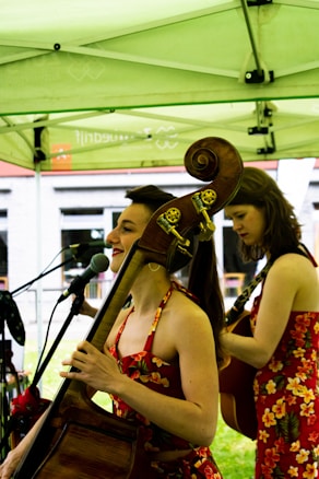 Two women wearing floral dresses perform music under a green canopy. One plays a double bass while the other plays a guitar. They stand in front of microphones, and natural light filters through the canopy.
