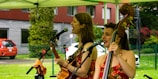 Two musicians perform outdoors under a canopy. One plays an acoustic guitar while the other plays a double bass, both singing into microphones. They wear matching floral dresses, and the background shows greenery and a parked car.