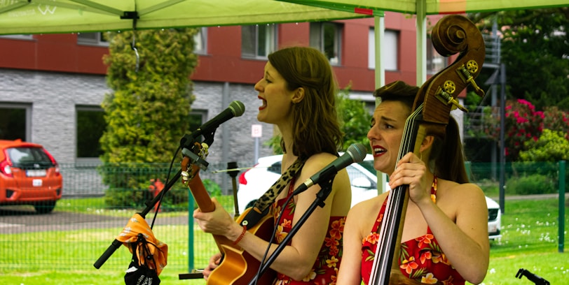 Two musicians perform outdoors under a canopy. One plays an acoustic guitar while the other plays a double bass, both singing into microphones. They wear matching floral dresses, and the background shows greenery and a parked car.