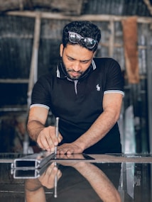 A man focuses intently while using a tool on a reflective surface. He wears a black polo shirt and has protective goggles on his head. The setting appears to be a workshop with industrial elements in the background.