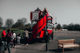 A red vintage double-decker bus is parked with several people boarding it. The bus has an open upper deck, and some individuals are seen climbing a staircase to reach it. The setting appears to be a park or open area with trees in the background and a bench on the right side of the image.