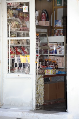 A small convenience store is visible through a partially open door. Shelves are stocked with various snacks, including bags of chips, candy, and boxes of goods. A refrigerator or display case is visible, suggesting the presence of perishable items. The store has a mix of domestic and imported products, with some shelves slightly cluttered.