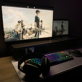 A dark, dimly lit computer setup featuring a large monitor and a laptop displaying an anime scene. The monitor shows several characters in action poses. A colorful RGB keyboard, a pair of headphones, and a mouse are placed on a desk in front.