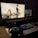 A dark, dimly lit computer setup featuring a large monitor and a laptop displaying an anime scene. The monitor shows several characters in action poses. A colorful RGB keyboard, a pair of headphones, and a mouse are placed on a desk in front.