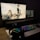 A dark, dimly lit computer setup featuring a large monitor and a laptop displaying an anime scene. The monitor shows several characters in action poses. A colorful RGB keyboard, a pair of headphones, and a mouse are placed on a desk in front.