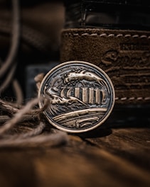 A metallic art piece with intricate engravings depicting waves and columns, encased in a round form. The background features textured brown leather and blurred twine in the foreground, adding depth and a rustic feel.