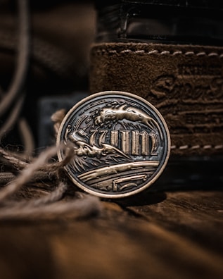 A metallic art piece with intricate engravings depicting waves and columns, encased in a round form. The background features textured brown leather and blurred twine in the foreground, adding depth and a rustic feel.