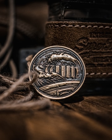 A metallic art piece with intricate engravings depicting waves and columns, encased in a round form. The background features textured brown leather and blurred twine in the foreground, adding depth and a rustic feel.