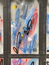 Graffiti art featuring elongated, stylized arrows in black, blue, and red, interspersed with small hearts and round yellow dots on a metal surface. Some splashes of white paint add texture.