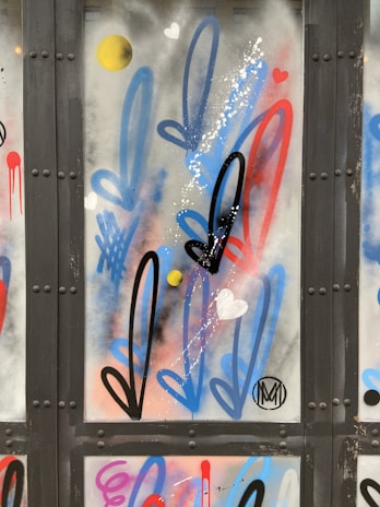 Graffiti art featuring elongated, stylized arrows in black, blue, and red, interspersed with small hearts and round yellow dots on a metal surface. Some splashes of white paint add texture.