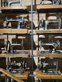 A collection of vintage sewing machines displayed on wooden shelves against a brick wall. The sewing machines are intricately designed with ornate patterns, and the shelves are supported by metal pipes. The lighting casts shadows on the wall, adding depth to the display.