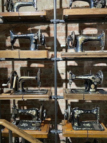 A collection of vintage sewing machines displayed on wooden shelves against a brick wall. The sewing machines are intricately designed with ornate patterns, and the shelves are supported by metal pipes. The lighting casts shadows on the wall, adding depth to the display.