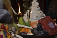 A lit brass oil lamp is surrounded by various ritualistic items, including incense sticks, a conch shell, flowers, and colorful cloth. The background shows a white tiered object decorated with mirrors.