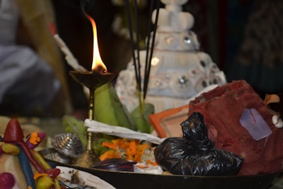 A lit brass oil lamp is surrounded by various ritualistic items, including incense sticks, a conch shell, flowers, and colorful cloth. The background shows a white tiered object decorated with mirrors.
