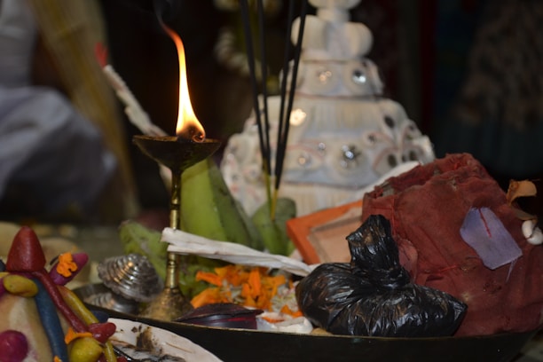 A lit brass oil lamp is surrounded by various ritualistic items, including incense sticks, a conch shell, flowers, and colorful cloth. The background shows a white tiered object decorated with mirrors.