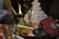 A lit brass oil lamp is surrounded by various ritualistic items, including incense sticks, a conch shell, flowers, and colorful cloth. The background shows a white tiered object decorated with mirrors.