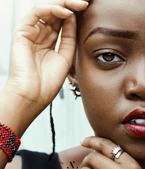 Close-up view of a person's face focusing on one eye and lips. One hand holds a section of hair while the other hand touches the chin. A beaded red and black bracelet and silver rings are visible. The makeup is bold with red lipstick and well-defined eyebrows.