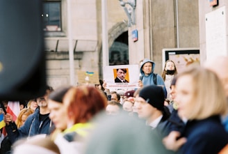 A protest scene with a crowd of people, some wearing hoodies and face masks, in front of a building. A person is holding up a sign featuring an image of a historical figure linked with propaganda text. Some flags are visible, and the crowd appears engaged in the demonstration.