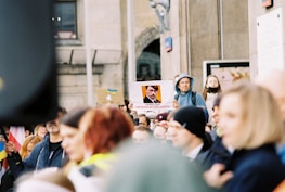 A protest scene with a crowd of people, some wearing hoodies and face masks, in front of a building. A person is holding up a sign featuring an image of a historical figure linked with propaganda text. Some flags are visible, and the crowd appears engaged in the demonstration.