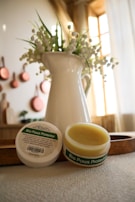 A white jug filled with delicate white flowers is set in a softly lit room. In front of the jug, there are two containers of 'Bio Paws Protector', with one open to reveal a yellow balm inside. The scene is calm and natural, suggesting a cozy home environment, with hints of a warm wooden and copper decor in the background.