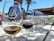 A selection of wine glasses filled with different types of wine is arranged on a wooden table, with a focus on a glass branded 'Die Mas'. The setting appears to be outdoors with a blurred background that includes a person and a palm tree.
