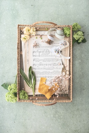 A wicker tray contains sheets of music accompanied by various decorative elements. There's a pair of scissors, a spool of ribbon, green foliage, flowers, and a piece of yellow cloth. The arrangement is artistic and neat, with a soft pastel background adding to the aesthetic appeal.