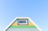 A brightly painted triangular house facade features pastel blue, green, and yellow horizontal stripes. It includes a small rectangular window with a white frame near the top. The facade is set against a clear blue sky, creating a fresh and vibrant appearance.