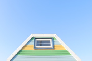 A brightly painted triangular house facade features pastel blue, green, and yellow horizontal stripes. It includes a small rectangular window with a white frame near the top. The facade is set against a clear blue sky, creating a fresh and vibrant appearance.