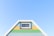 A brightly painted triangular house facade features pastel blue, green, and yellow horizontal stripes. It includes a small rectangular window with a white frame near the top. The facade is set against a clear blue sky, creating a fresh and vibrant appearance.