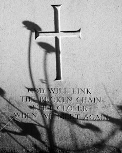 A stone surface features a carved cross with an inscription beneath it. Shadows of plants are cast across the surface, adding contrast to the image. The inscription reads, 'God will link the broken chain still closer when we meet again.'