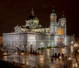 An illuminated cathedral stands majestically against a night sky, showcasing a blend of neo-classical and baroque architecture. The building is adorned with domes, towers, and intricate details, surrounded by a wet cobblestone plaza reflecting the light from surrounding streetlamps.