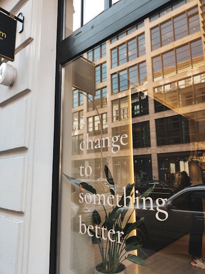 A storefront window reflects a city street with multiple buildings and parked cars. The words 'change to something better' are printed on the window. Inside, a green plant is visible, adding a natural element to the urban setting.