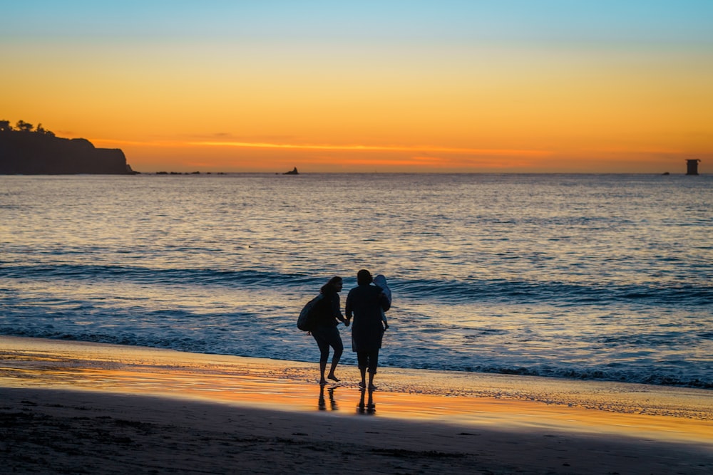 a couple of people walk on the beach with surfboards