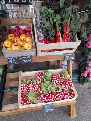A display of fresh produce at a market with boxes filled with colorful fruits and vegetables. There are golden and red apples, vibrant red and green rainbow chard, and bunches of bright red breakfast radishes with green leaves. Each type of produce is labeled with small slate signs showing their names and prices.