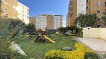 A playground with a wooden play structure featuring slides and climbing areas, surrounded by well-maintained grass and yellow flowering bushes. Nearby are benches for seating. The area is enclosed by residential buildings with multi-storey apartments. Trees and additional greenery line the edges of the playground, and there is a small soccer goal on a paved area.
